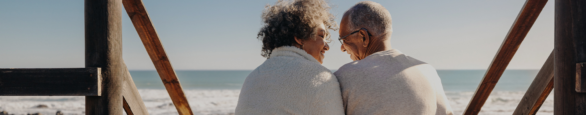 An older couple smile at each other while sitting on a staircase at the beach looking at the ocean.