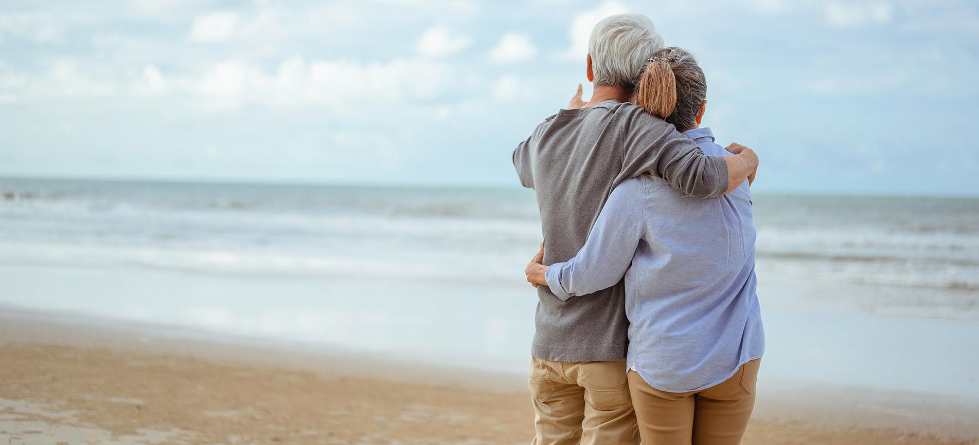 An older couple in an embrace standing on the beach while looking out and pointing to the sea.