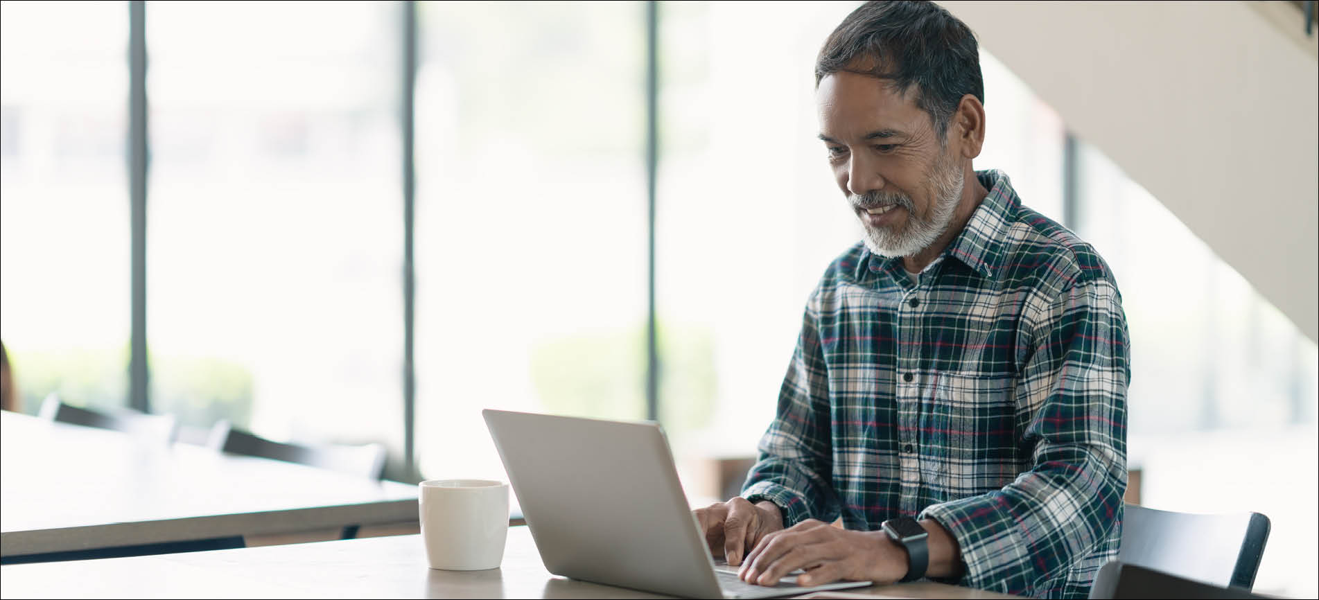 A man with a grey beard and wearing a plaid shirt smiles while using a laptop in a bright, modern room. A coffee cup sits nearby on the wooden table.